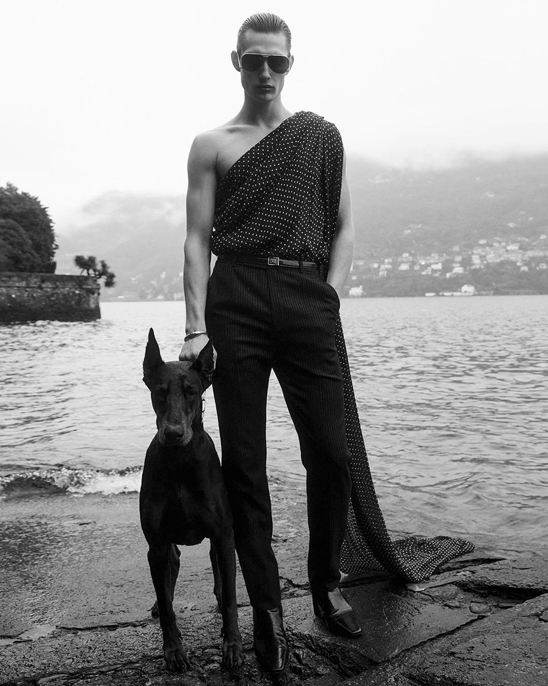 Monochrome fashion shot of a man in an off-the-shoulder polka dot top and pinstripe pants standing with a Doberman on a rocky shoreline.
