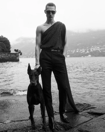 Monochrome fashion shot of a man in an off-the-shoulder polka dot top and pinstripe pants standing with a Doberman on a rocky shoreline.