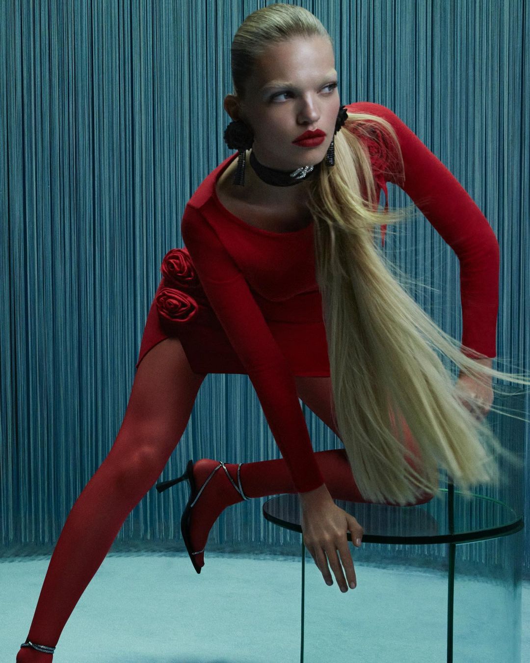 A blonde fashion model poses indoors, wearing a red dress, red stockings, and black high-heeled shoes, beside a glass table against a blue curtain backdrop.