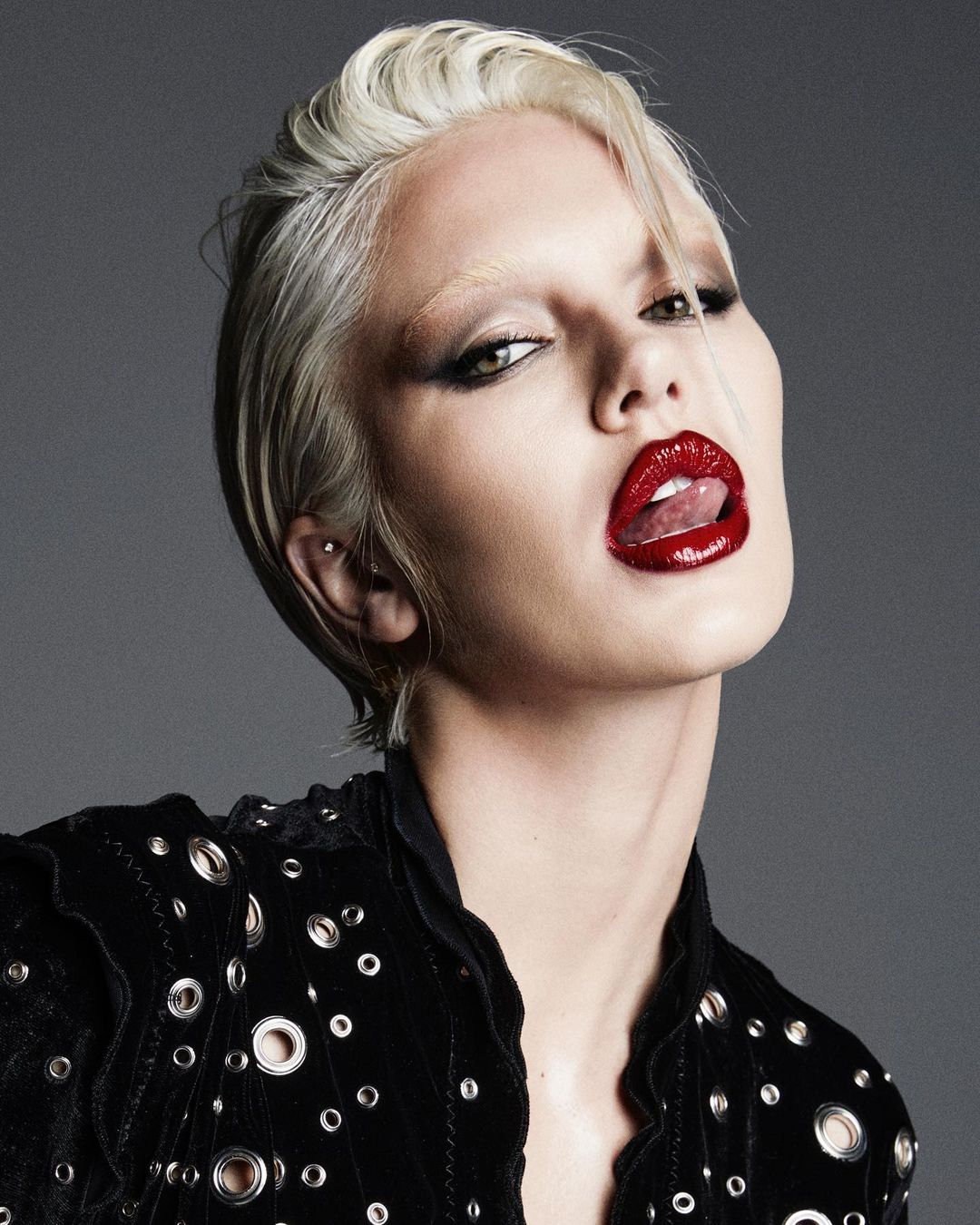 Close-up portrait of a model with platinum blond hair, dark eye makeup, and red lipstick sticking out her tongue against a gray background.