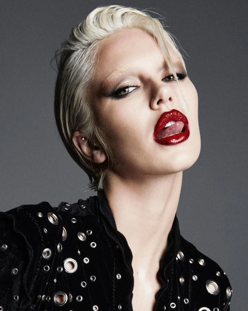 Close-up portrait of a model with platinum blond hair, dark eye makeup, and red lipstick sticking out her tongue against a gray background.