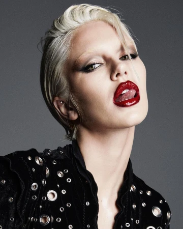 Close-up portrait of a model with platinum blond hair, dark eye makeup, and red lipstick sticking out her tongue against a gray background.