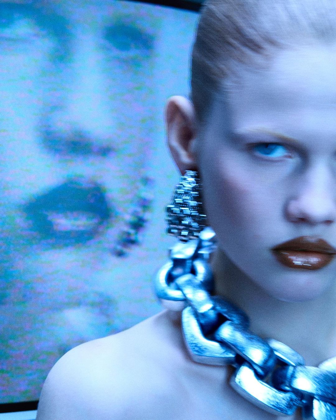 Close-up of a woman with dark lipstick and silver chain jewelry in front of a blurry TV showing a person's face.