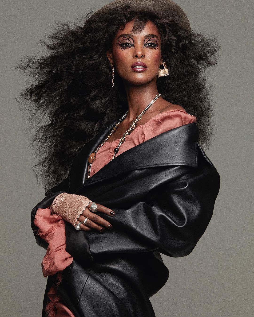 Woman with voluminous curly hair wearing a black leather coat and pink top, photographed against a grey background.