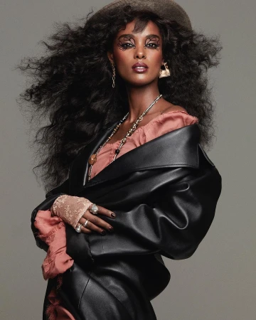 Woman with voluminous curly hair wearing a black leather coat and pink top, photographed against a grey background.