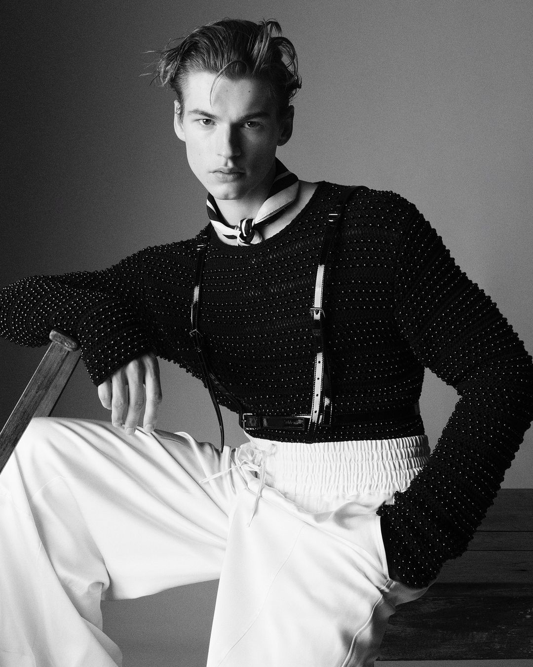 Black and white fashion shot of an adult male wearing a textured black top, white pants, and suspenders, posed in a studio setting.