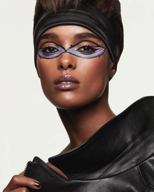 Close-up of a dark-skinned woman with painted eye makeup, black headband, and glossy lips, wearing a black leather jacket on a white background.