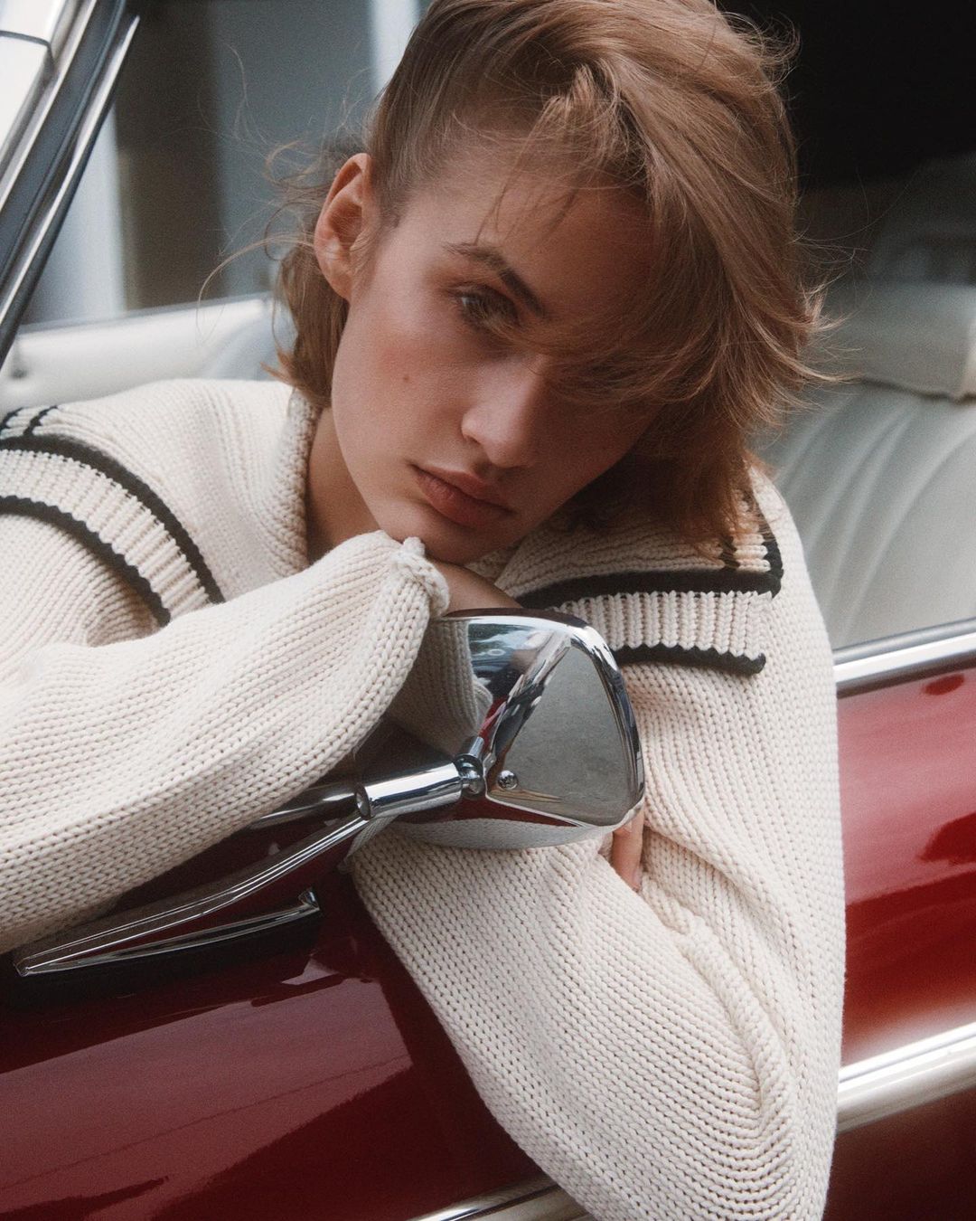 Portrait of a young adult female with short blonde hair in a white knit sweater posing near a red car, with the car's side mirror visible.