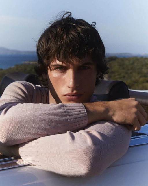 Close-up of a brunette male model in a light pink sweater leaning against a car with an ocean and island background on a clear day.