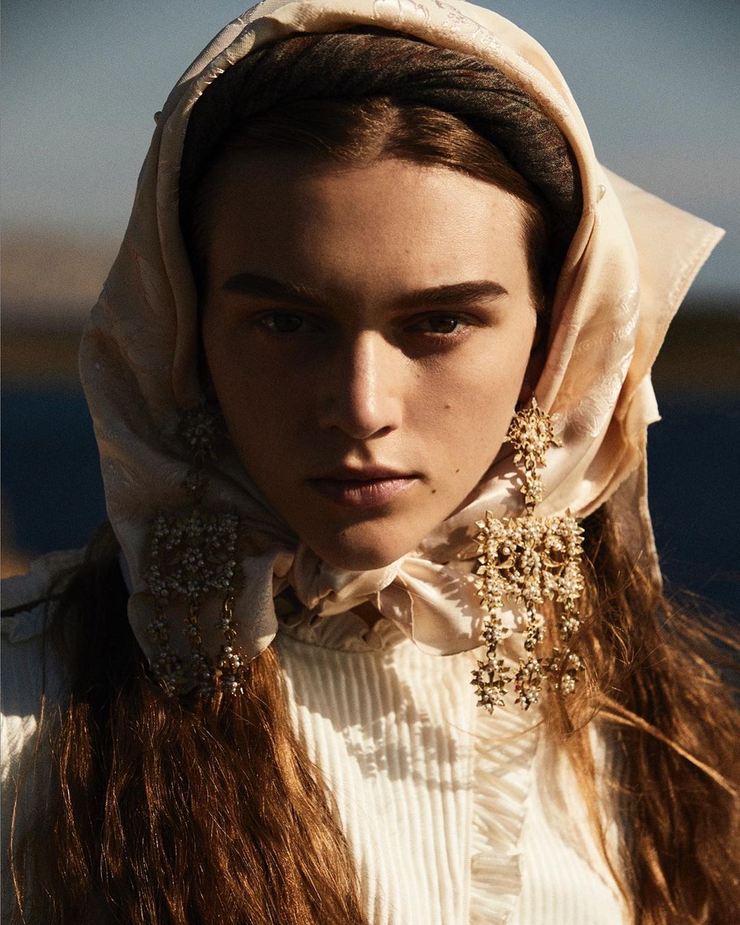 Close-up portrait of a woman wearing a cream silk headscarf and ornate gold statement earrings by photographer Alvaro Beamud Cortes.