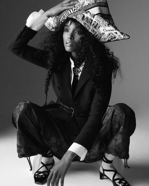 Black and white portrait of a woman in a blazer, printed pants, and strappy heels, holding a printed Christian Dior hat.