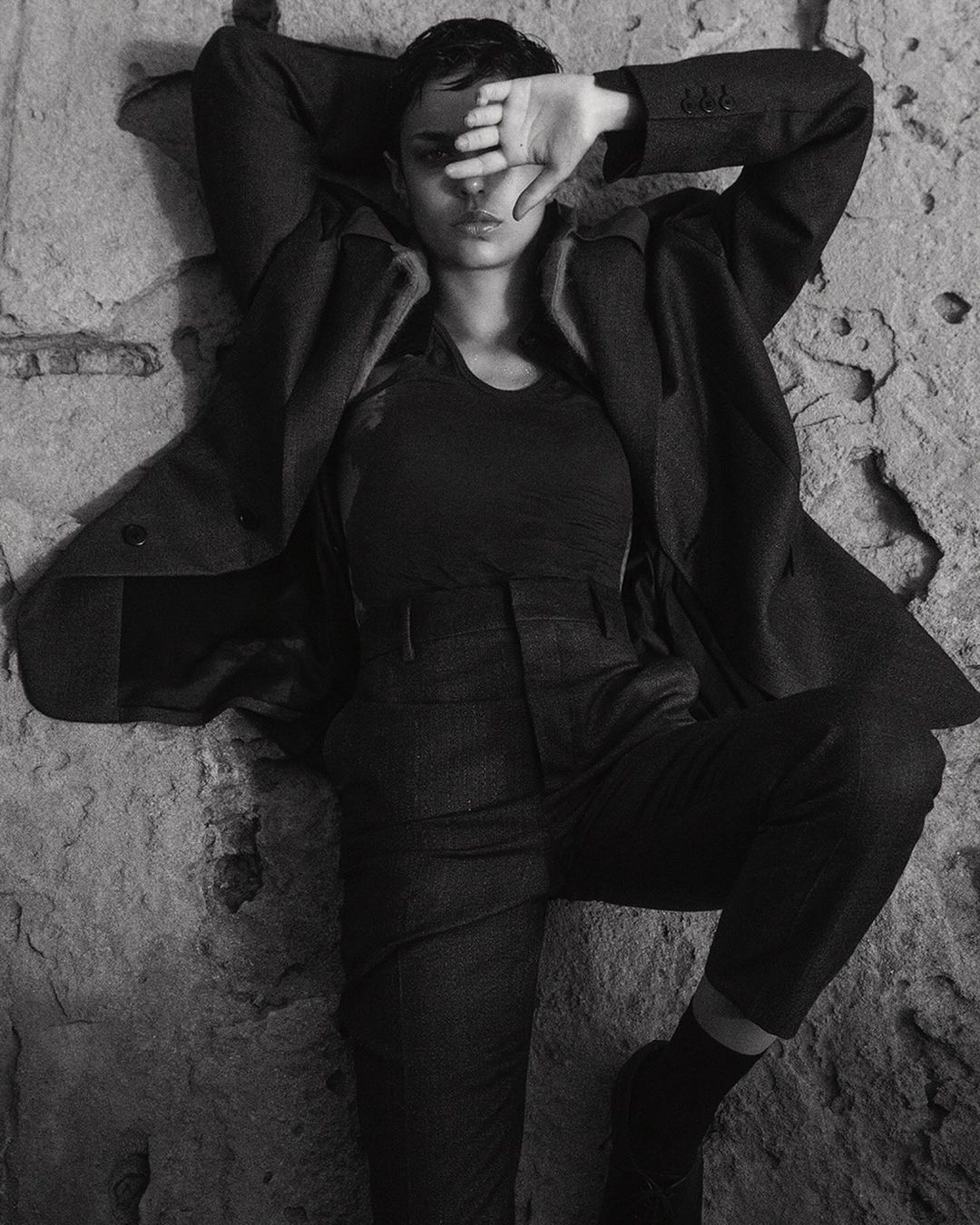 Model with short dark hair lying against a rough textured stone wall wearing a tailored jacket and trousers.