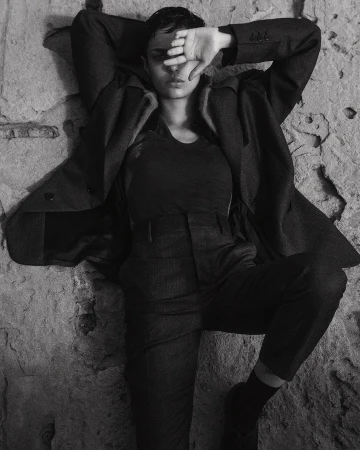 Model with short dark hair lying against a rough textured stone wall wearing a tailored jacket and trousers.