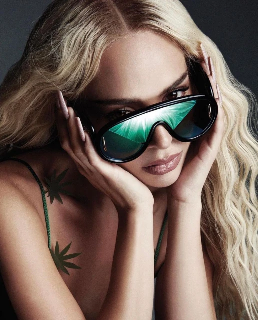 Close-up of a blonde adult female wearing dark sunglasses and a green top with leaf accents against a dark grey background.