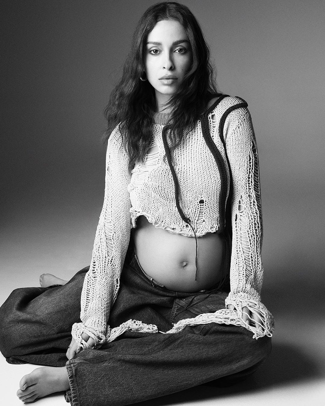 Black and white portrait of a pregnant woman with long dark hair, wearing a knitted top and jeans, seated in a photo studio.