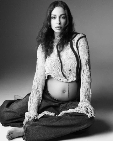 Black and white portrait of a pregnant woman with long dark hair, wearing a knitted top and jeans, seated in a photo studio.