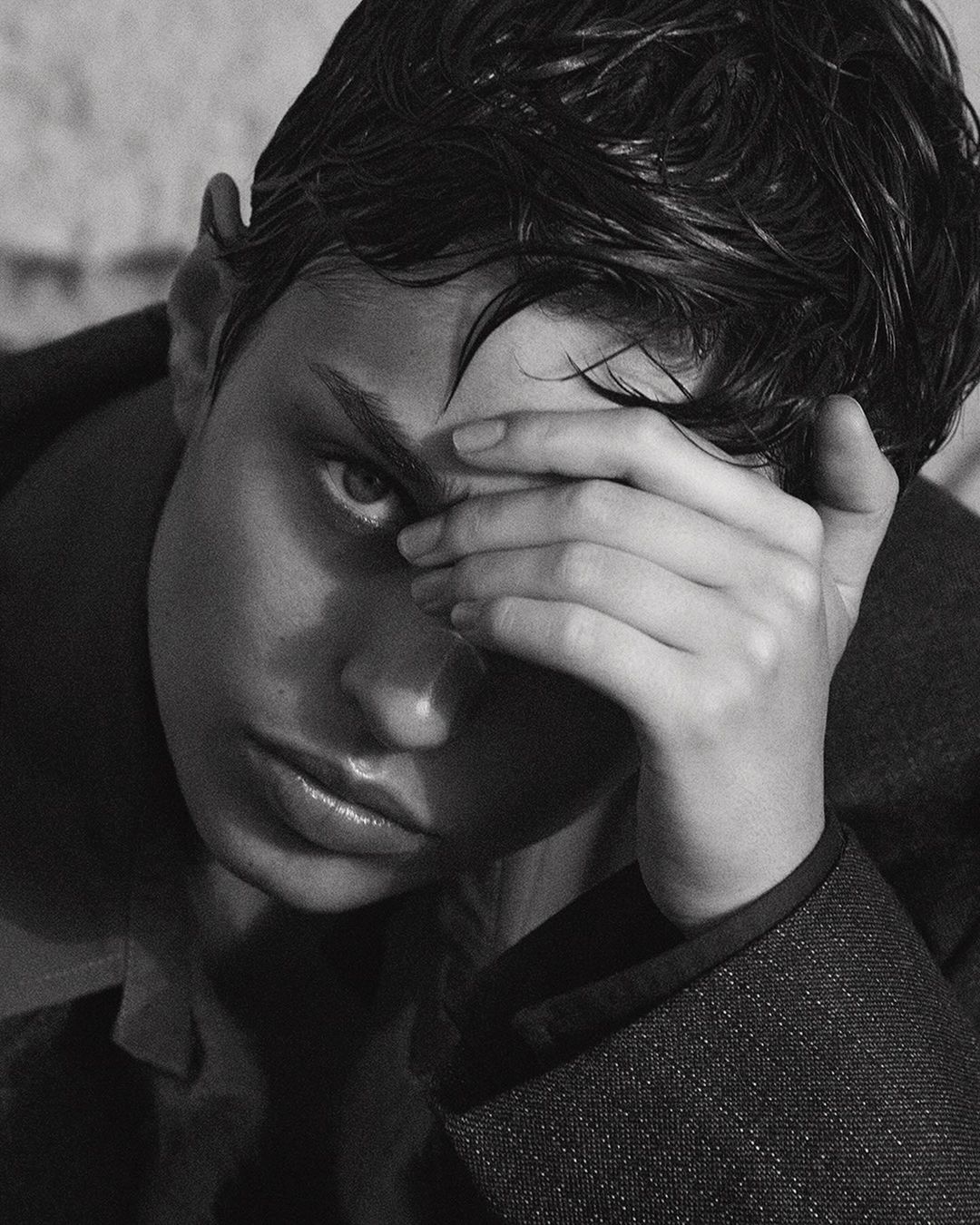 Close-up black and white portrait of a model with wet dark hair holding a hand to their forehead.