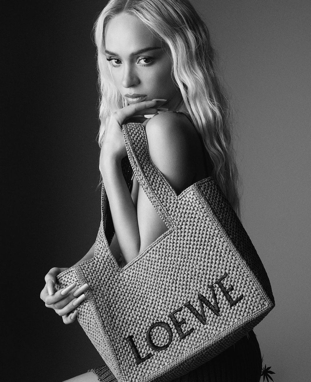 Black and white portrait of a woman with blonde wavy hair, posing with a raffia tote bag with the word 'LOEWE' displayed.