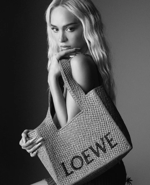Black and white portrait of a woman with blonde wavy hair, posing with a raffia tote bag with the word 'LOEWE' displayed.