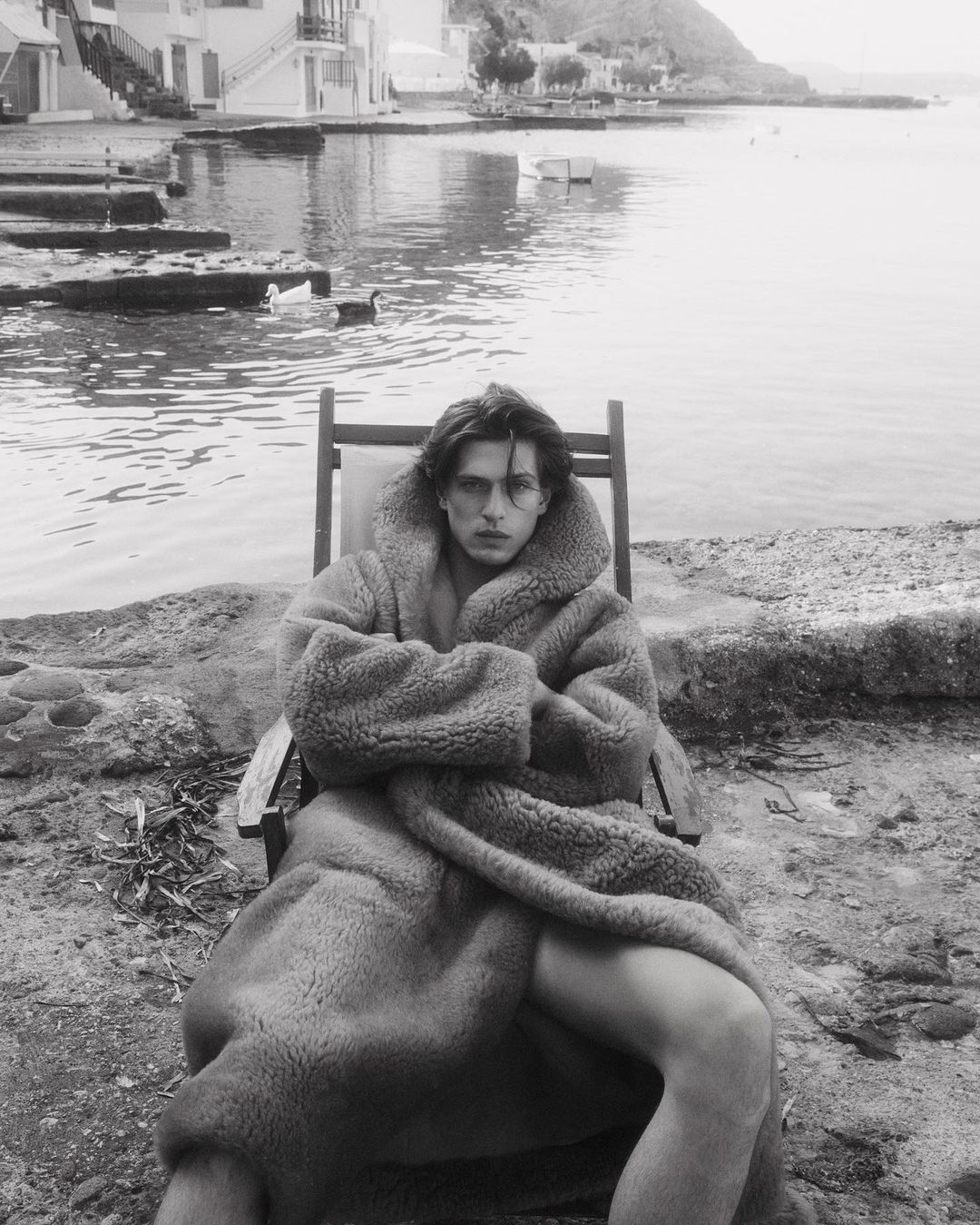 Monochrome portrait of a man in a heavy plush hooded robe seated in a deckchair by the water.