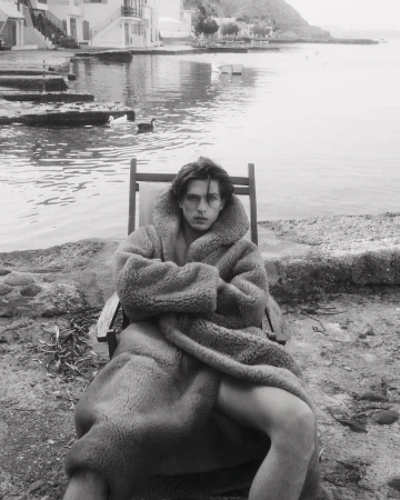 Monochrome portrait of a man in a heavy plush hooded robe seated in a deckchair by the water.