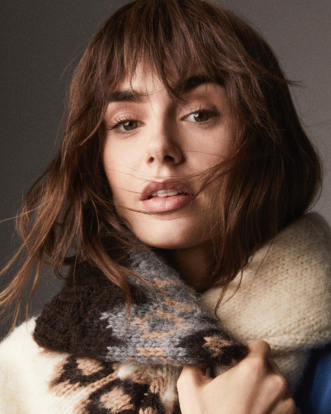 Close-up portrait of a woman with brown hair and bangs wearing a chunky patterned knit scarf and cream sweater.