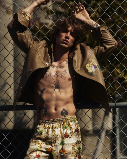 A shirtless man poses in a tan jacket and patterned shorts against a chain-link fence, showcasing tattoos, style, and urban fashion.