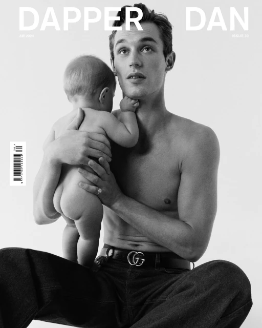 Black and white magazine cover featuring a shirtless man holding a baby, both looking upwards. The man is wearing jeans and a Gucci belt.