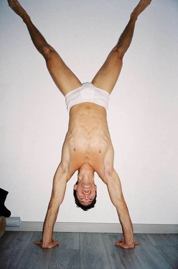 Adult male performs a handstand wearing briefs. The man is smiling and looking towards the camera in an indoor environment.