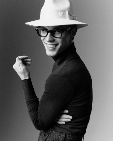 Man wearing white fedora, black eyeglasses, and dark turtleneck smiling over his shoulder against a grey background.