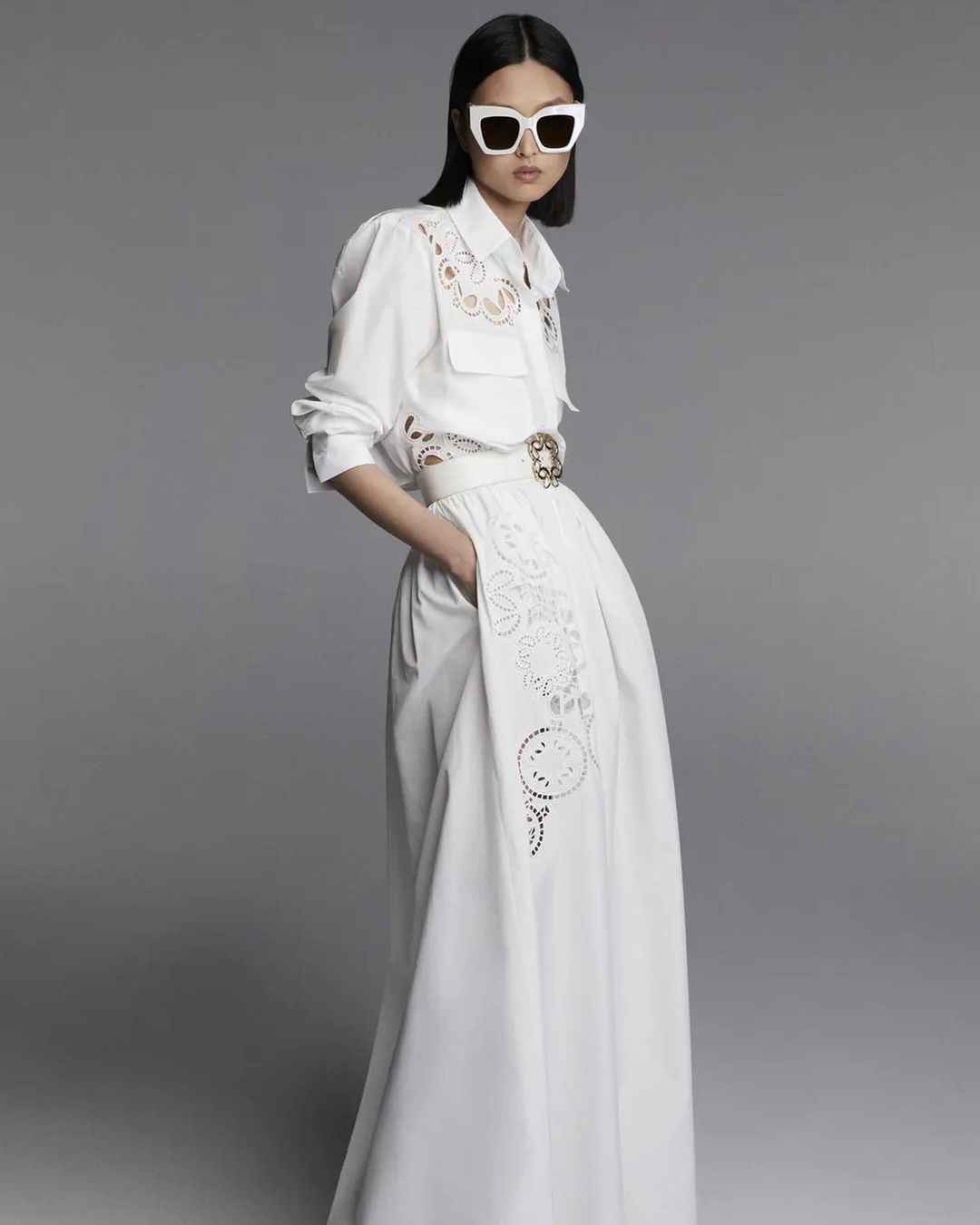 An Asian woman stands in a white long-sleeved dress with a white belt and sunglasses, posing against a neutral grey backdrop in a fashion editorial.