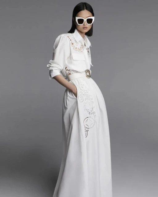 An Asian woman stands in a white long-sleeved dress with a white belt and sunglasses, posing against a neutral grey backdrop in a fashion editorial.