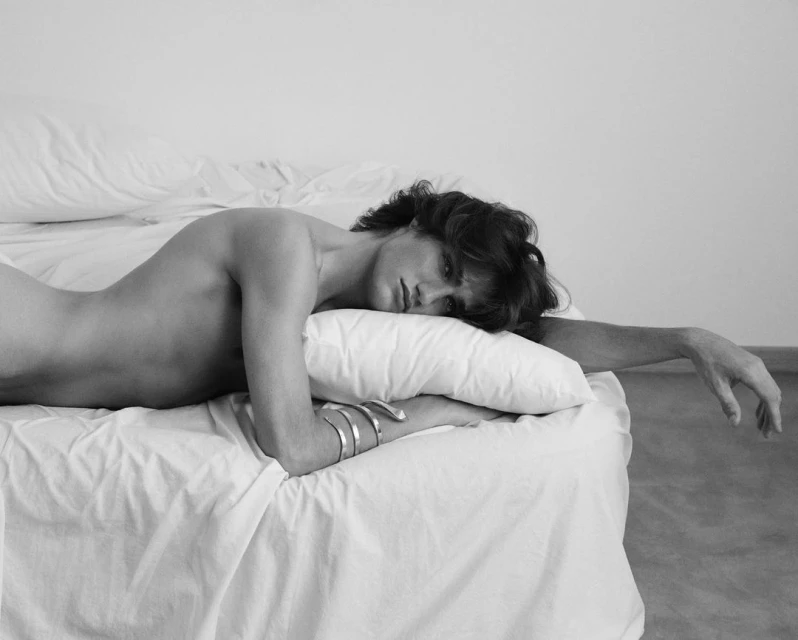 Black and white photo of a young adult male lying on a white bed with a white pillow, showing his bare back, arm, and dark hair.