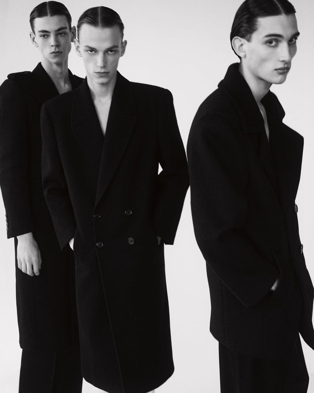 Three male fashion models in black wool overcoats and slicked-back hair stand indoors against a light background in a studio photoshoot.