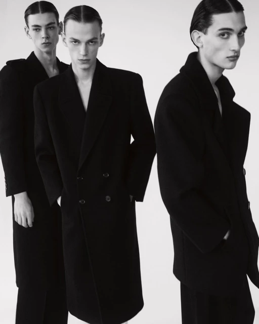 Three male fashion models in black wool overcoats and slicked-back hair stand indoors against a light background in a studio photoshoot.