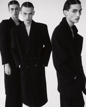 Three male fashion models in black wool overcoats and slicked-back hair stand indoors against a light background in a studio photoshoot.