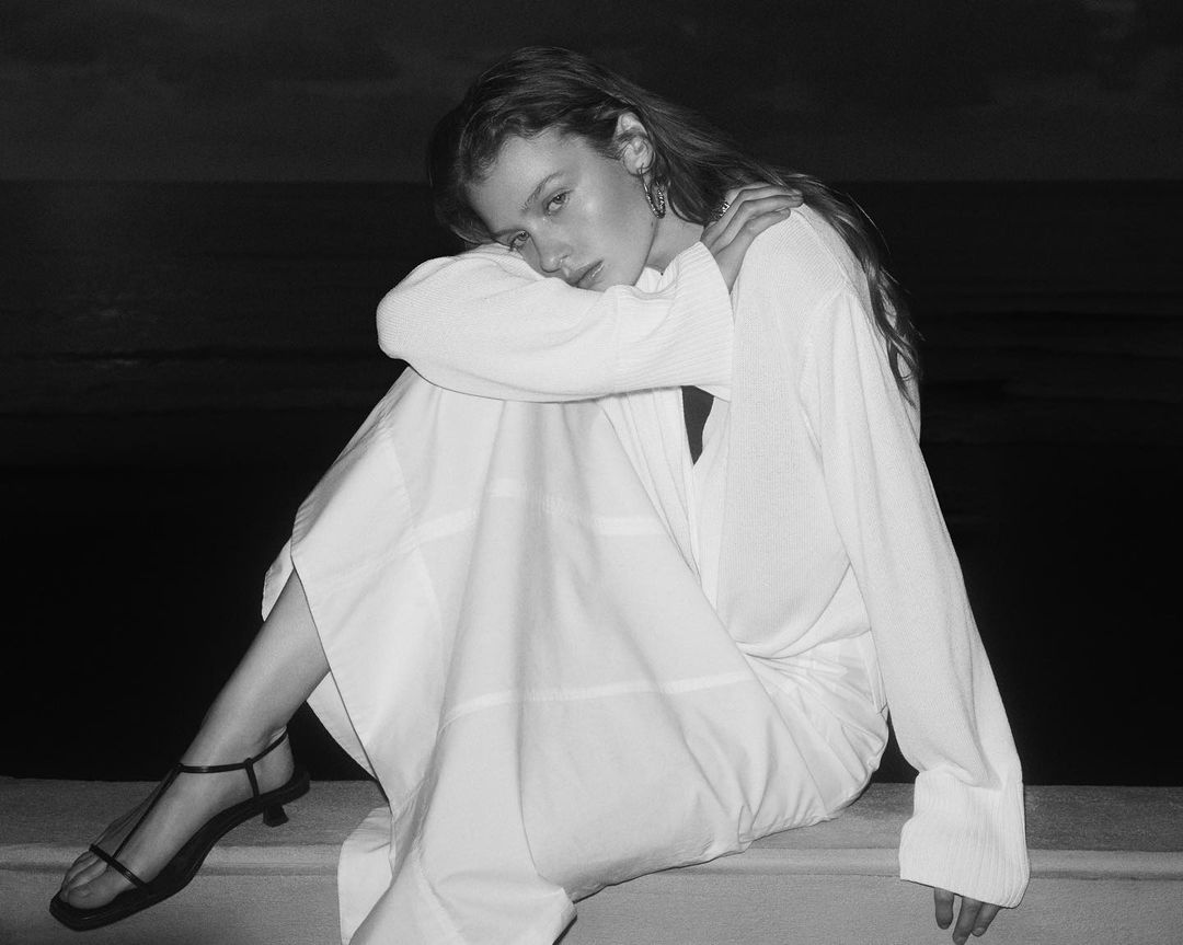 Black and white photo of a fair-skinned female model in a white dress and black strappy heels sitting on a ledge at twilight.