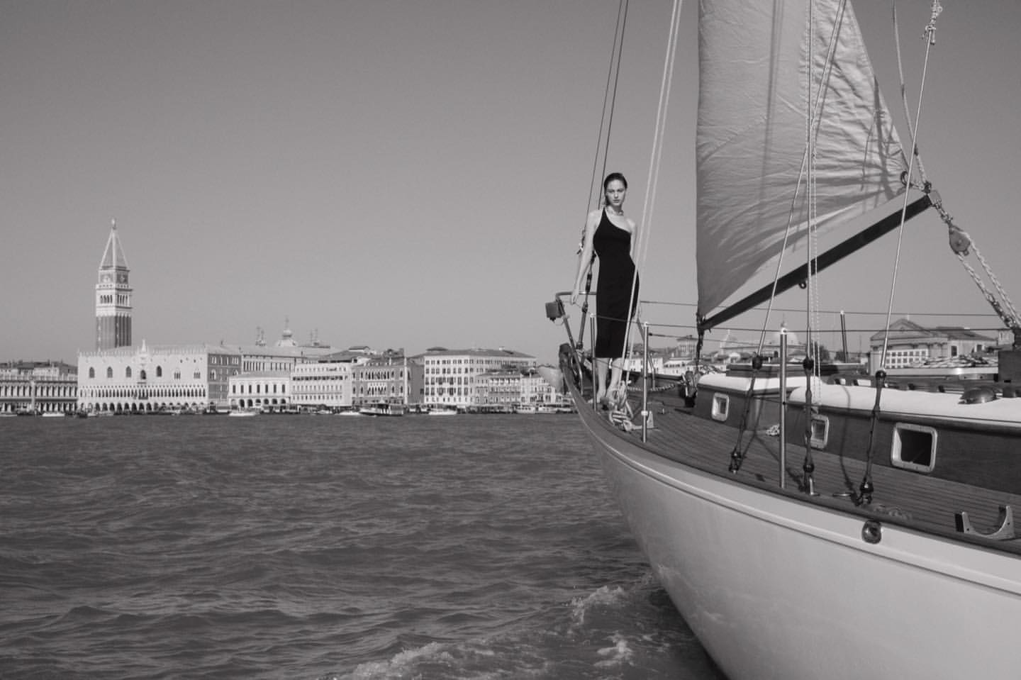 A woman in a dress stands on a sailboat with the Venice skyline in the background on a bright day in a monochrome photograph.