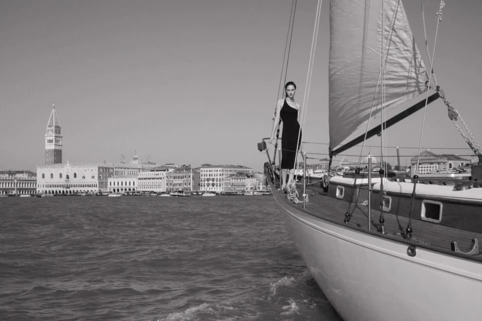 A woman in a dress stands on a sailboat with the Venice skyline in the background on a bright day in a monochrome photograph.