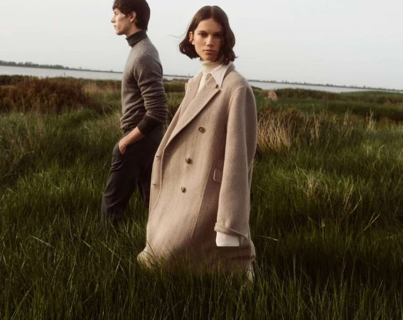 Two models standing in a tall green grassy field outdoors with a body of water in the background, photographed by Alvaro Beamud Cortes.