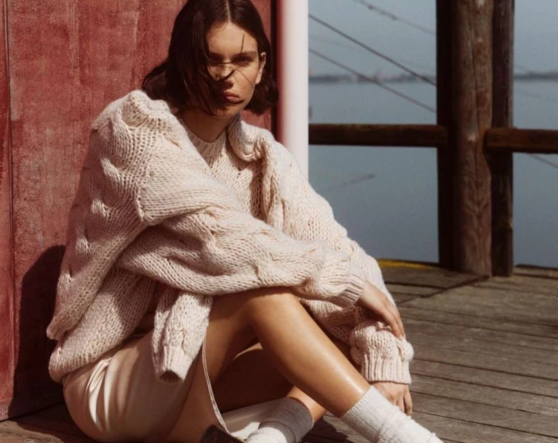 Brunette woman in chunky knit sweater, mini dress, and socks, sitting on wooden planks near a body of water on a sunny day.