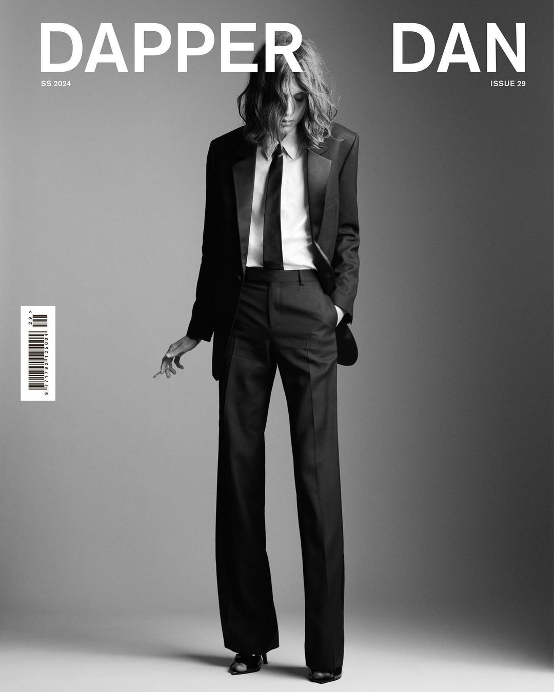 Black and white magazine cover featuring a person in a suit, tie, and long hair, standing against a gray background.