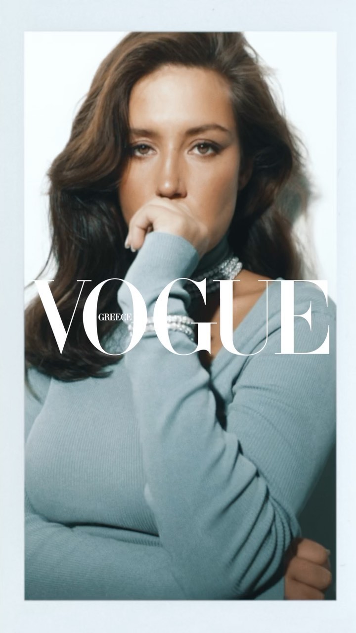 Close-up of woman in blue top, hand to face, against white backdrop, with 'Vogue Greece' logo overlay, showcasing fashion and editorial style.