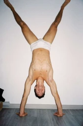 Man doing a handstand wearing white briefs against a white wall with grey wooden flooring by Alvaro Beamud Cortes
