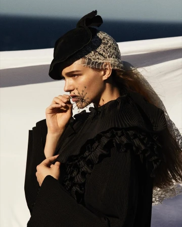 Model in black ruffled top holding a delicate plant stem to her mouth, wearing a structured hat and lace head covering against an ocean background.