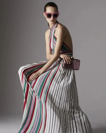 Model in pink sunglasses wearing a striped halter dress holding a clutch against a grey background