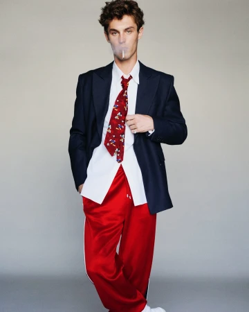 Young man in dark blazer, white shirt, red patterned tie, and red track pants standing against a grey backdrop