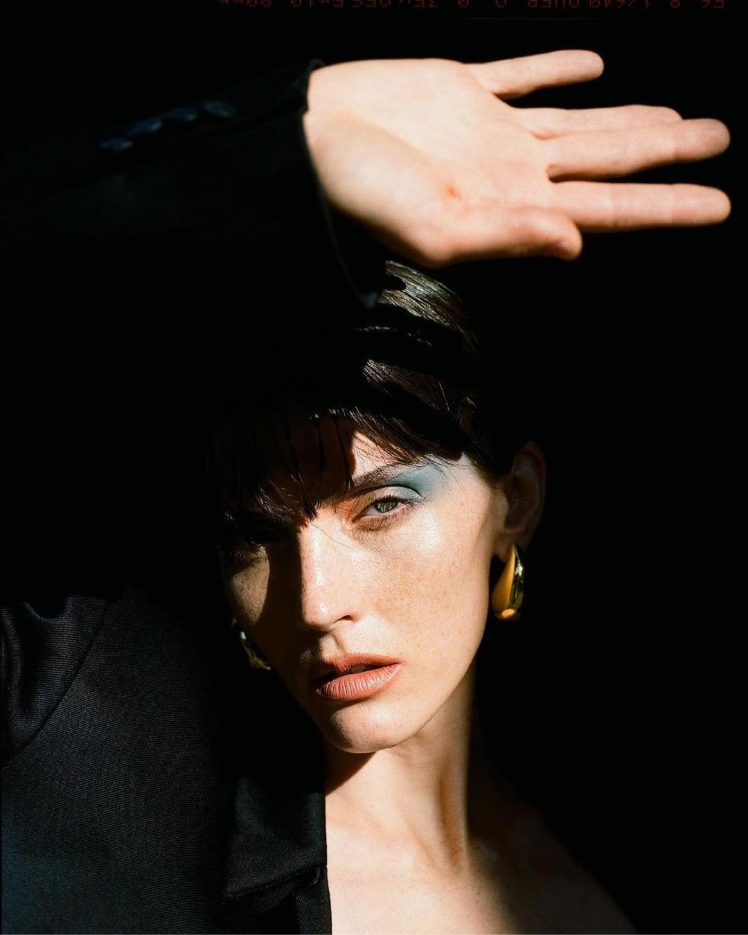 Portrait of a woman with blue eyeshadow, gold earring, and black top shielding her face from sunlight with her hand.