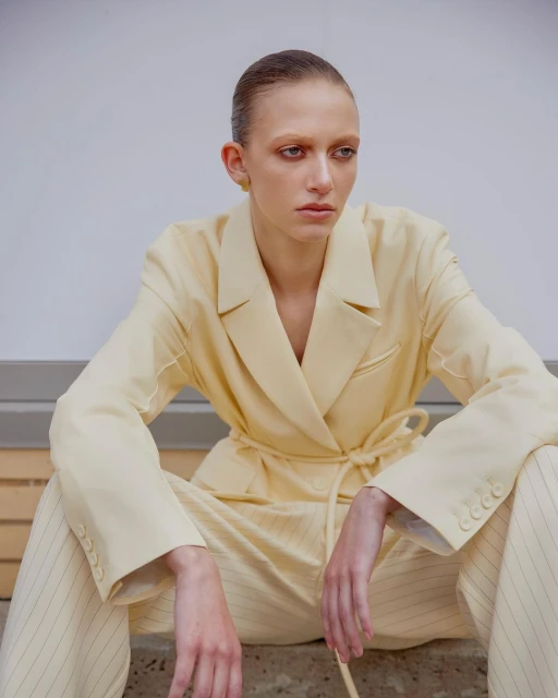 A woman in a yellow suit sits in an indoor setting with a white background. She looks off to the side while seated on a concrete step.