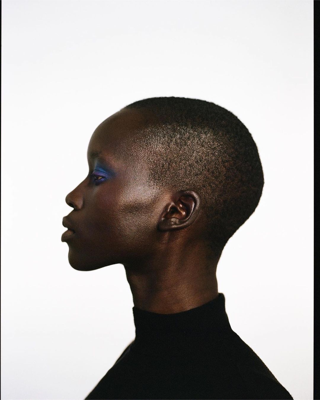 Profile portrait of a Black woman with short hair wearing a black turtleneck, showcasing makeup against a plain white background.